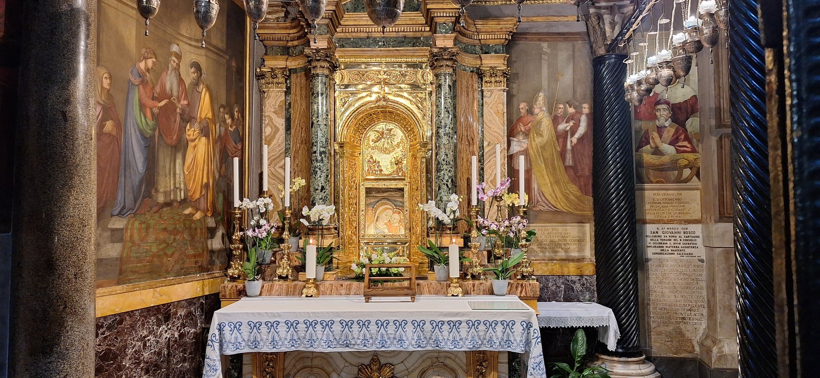 Altar e imagen de la Virgen del Buen Consejo, rodeados de pinturas, flores y columnas.